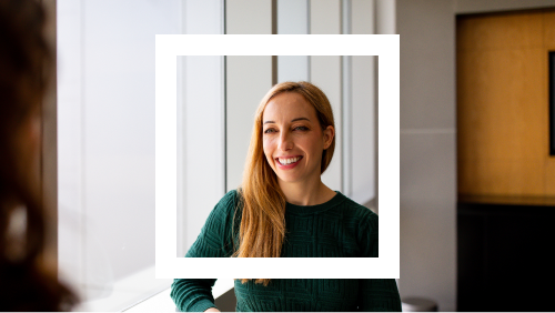 Portrait of a smiling woman with long straight blonde hair, wearing a dark green textured blouse, posed in a bright room.