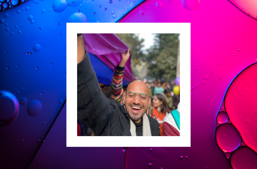 A joyful person wearing glasses and a dark jacket smiles widely while raising his arm during a colorful outdoor event. Behind him, people are gathered, holding vibrant banners and balloons. The image is framed by a white border and set against a vibrant blue and pink abstract background with bubble-like patterns. 