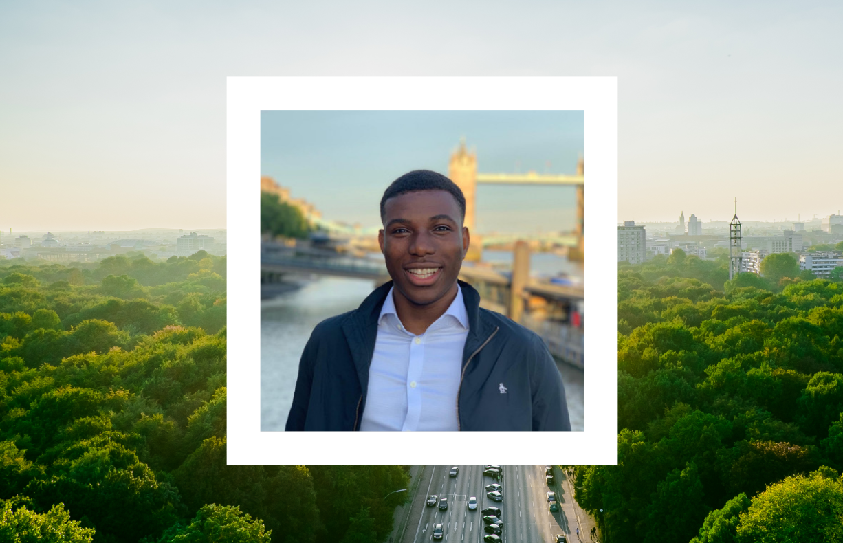 A smiling man in a dark jacket and white shirt stands outdoors with a bridge and river in the background. The image is framed within a white square overlay against a scenic view of a green forest and a city skyline at sunset.