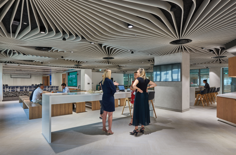 A communal workspace with people engaging in conversation, surrounded by open-plan desks and the same distinctive ceiling design.