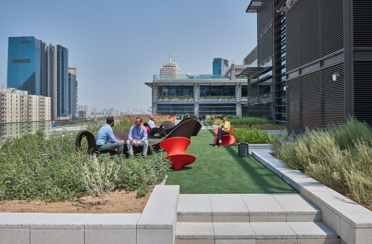 An outdoor seating area with vibrant red chairs, greenery, and a cityscape in the background, offering a relaxed meeting space.