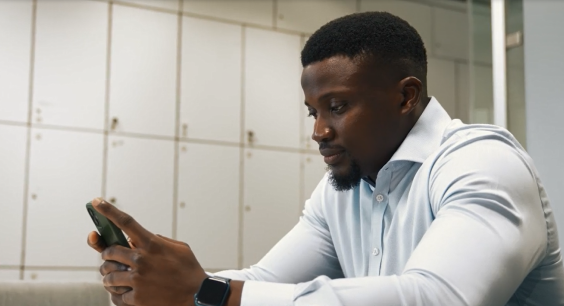 A man in a light blue dress shirt sits at a table, focused on his smartphone. He has a short, neat haircut and a closely trimmed beard. He is wearing a smartwatch on his left wrist. The background features a wall with a grid of white storage lockers, suggesting a modern office or coworking space. The atmosphere is professional, with the man appearing engaged and attentive to the information on his phone.