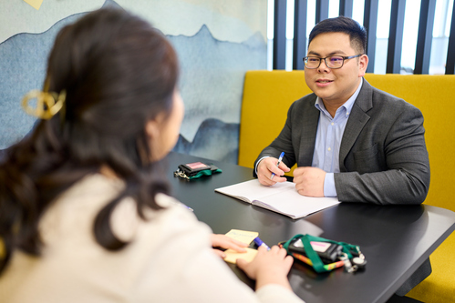 Portrait of a smiling man with short black hair, wearing a light blue checkered shirt, seated in a modern office setting with glass partitions and a concrete wall in the background.