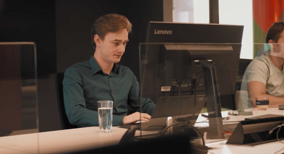 A young man with light brown hair is sitting at a desk, focused on his work at a Lenovo desktop computer. He is wearing a dark green shirt and has a glass of water beside him. The background shows a modern office setting with another person working at a nearby desk, partially visible.