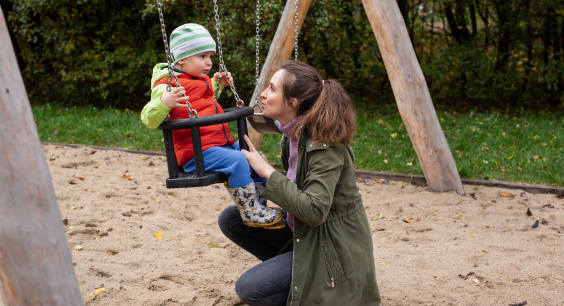 A woman kneels beside a young child sitting in a swing at a playground. The child, dressed in a red vest, blue pants, green jacket, and striped beanie, looks ahead while holding onto the swing chains. The woman, wearing a green coat, gently holds the swing and looks at the child with a caring expression. The playground is set in a wooded area with a sand-covered ground and wooden swing frame.