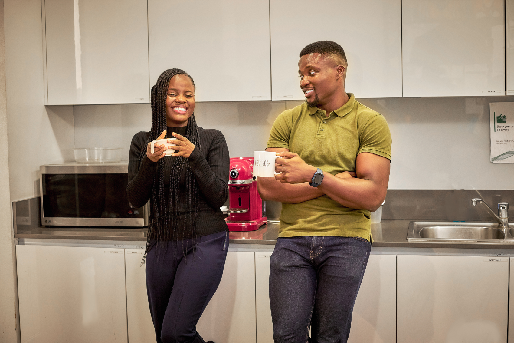 Two colleagues are sharing a light moment in a modern office kitchen. A woman on the left, with long braids and wearing a dark knit top with slim-fitting trousers, is smiling broadly, holding a small cup with both hands. A man on the right, in a fitted olive-green polo shirt and dark jeans, is holding a white mug, looking at the woman with an amused expression. They're standing next to a bright pink kitchen appliance, possibly a coffee machine, with a microwave above and a dishwasher below.