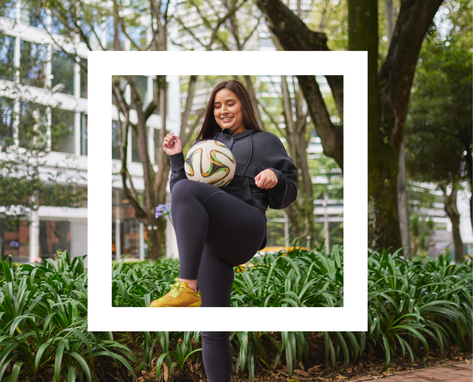 A woman with long hair, wearing a dark tracksuit and yellow sneakers, is outdoors in an urban park setting. She is smiling and playfully holding a soccer ball with her leg, surrounded by greenery and trees with buildings in the distance.