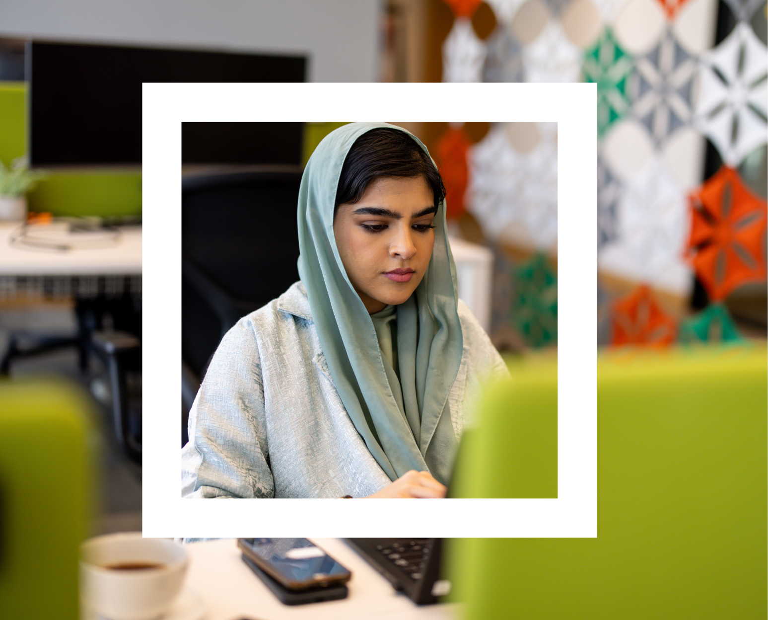 A person is sitting at a desk in an office environment, working on a laptop. The person is wearing a light green hijab and a grey top. In the background, there's a white monitor, some colorful geometric wall art, and part of a green office chair is visible in the foreground. 