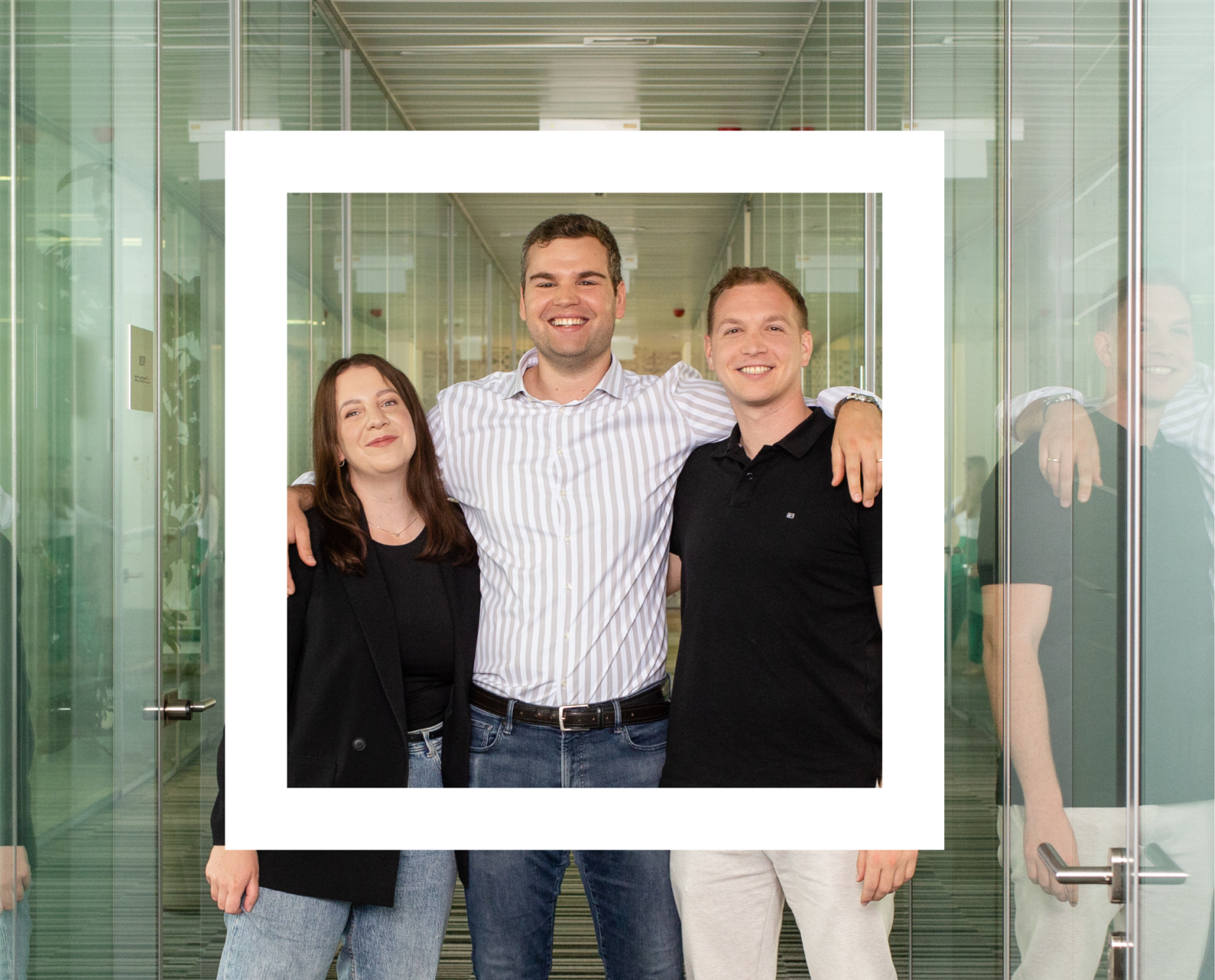 Three individuals standing closely together, posing for a photo in an office setting with transparent glass doors in the background. The person in the center is wearing a white and gray striped shirt with blue jeans, flanked on either side by individuals wearing black tops. 