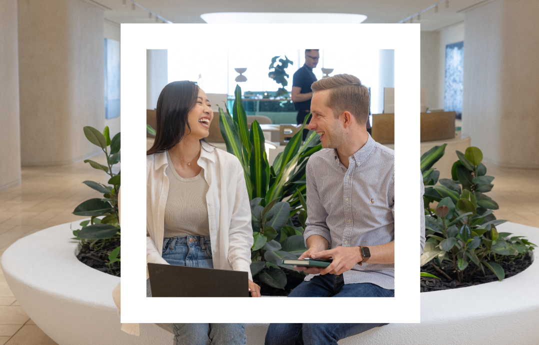 A Polaroid-style photograph features two people seated next to each other in a modern, indoor space with neutral tones and contemporary decor. They are sitting on the edge of a circular planter filled with lush green plants. The woman on the left, wearing a white shirt and jeans, is laughing while looking at the man. The man on the right, wearing a light blue striped shirt, is smiling back at her while holding a tablet. The background shows another person standing and working in the spacious, well-lit room, adding to the professional and friendly atmosphere.