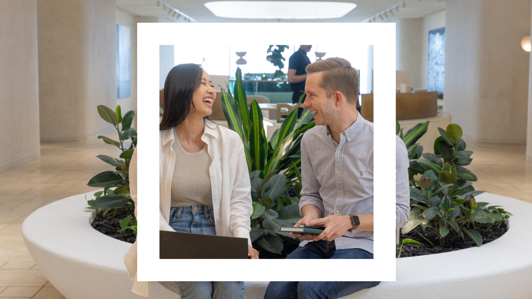 A woman and a man sit on a round bench surrounded by plants in a modern indoor space. They are laughing and engaging in a conversation, with the woman holding a laptop and the man holding a notebook. The image is framed within a white rectangular overlay.