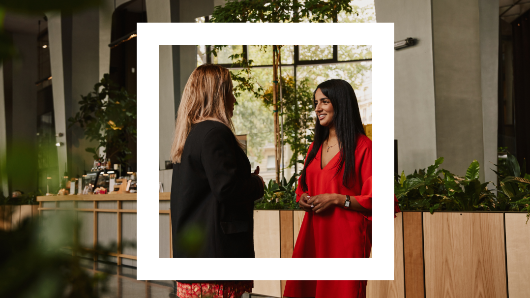  A professional image capturing a moment between two women engaged in conversation in a modern office cafeteria. The woman on the right, dressed in a vibrant red dress, is smiling and talking to the other woman, who is wearing a black business suit and is seen from the back. The setting is well-lit and features contemporary wooden furniture with lush green plants around, creating a relaxed yet professional atmosphere.