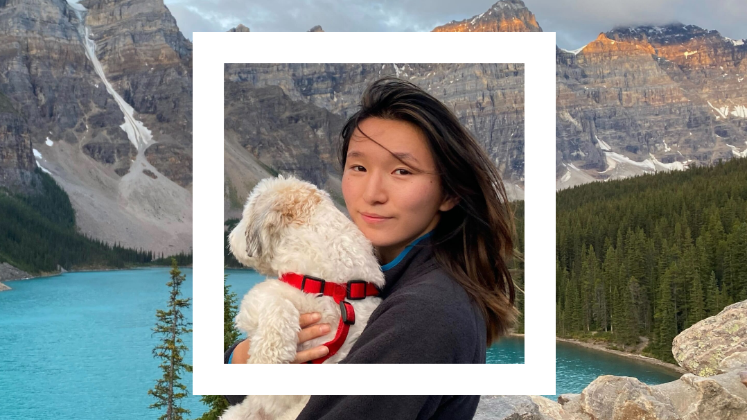 A young woman with long dark hair stands outdoors, holding a small white dog wearing a red harness. They are set against a breathtaking backdrop of turquoise blue water, evergreen trees, and towering rocky mountains, partially covered with patches of snow. The image is framed by a white Polaroid-style border, adding a nostalgic touch to the scenic, natural setting. The woman gazes directly at the camera, her expression calm and content. 