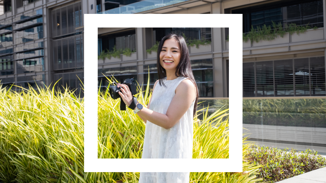 A woman in a white sleeveless dress stands outdoors smiling while holding a camera. She has long dark hair and is surrounded by tall green plants. A white rectangular border frames the central part of the image, emphasizing her against the urban backdrop with buildings and windows.