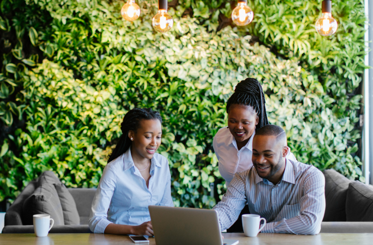 two women and on man working on a laptop infront of a large plant