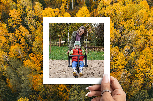  A person is holding a polaroid-style photo of a young child on a swing with an adult behind them, both smiling. The background of the photo shows a green grassy area and some trees. This polaroid photo is being held up against a background of a dense forest with vibrant autumn foliage, predominantly yellow and some green.