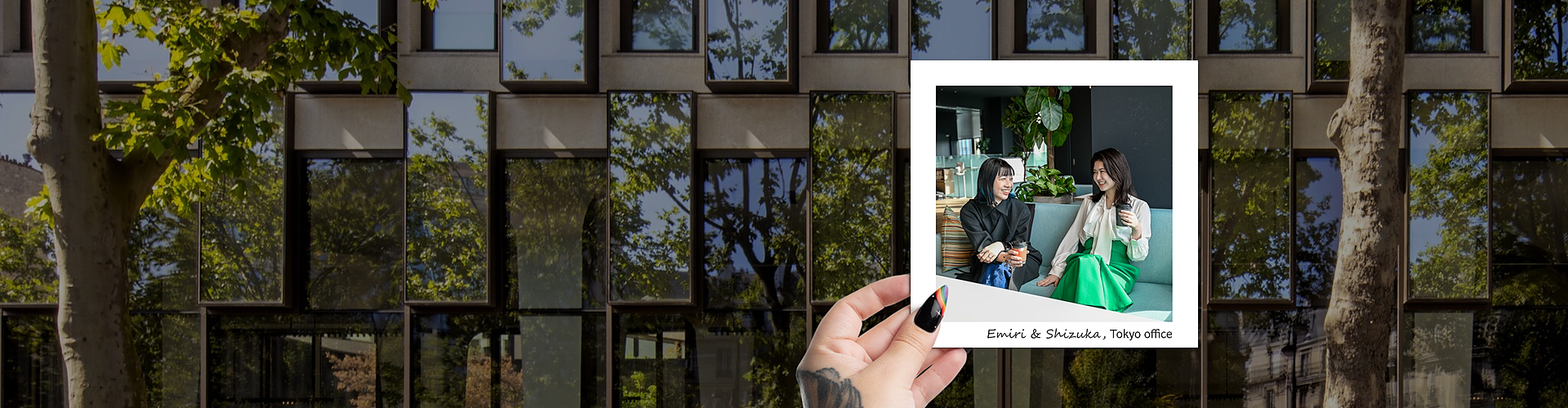 A hand with colorful nail art is holding a polaroid photo against the backdrop of a modern building with large windows reflecting green trees. The polaroid photo features two women, Emiri and Shizuka, sitting and smiling at each other in an office setting. One woman is wearing a black jacket, and the other is in a green skirt and white top. The text on the polaroid reads "Emiri & Shizuka, Tokyo office."