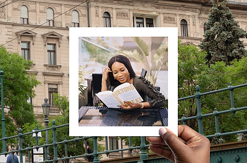 A creative photo depicts a hand holding a Polaroid of a young woman reading a book titled "Beyond Great" at an outdoor café. The background outside the Polaroid shows an urban park setting with lush trees and historical architecture, contrasting with the intimate, focused scene in the photo. The woman is stylishly dressed and appears engrossed in her reading.