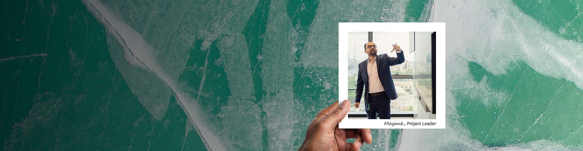 This panoramic banner displays a hand holding a photograph against a textured backdrop of cracked ice on a frozen lake, with shades of green and white enhancing the natural, rugged beauty. The photograph features Mayank, a project leader, actively engaging in a presentation or discussion in a modern office environment. He is using his hand to emphasize a point, and his focused expression suggests a serious business context. 