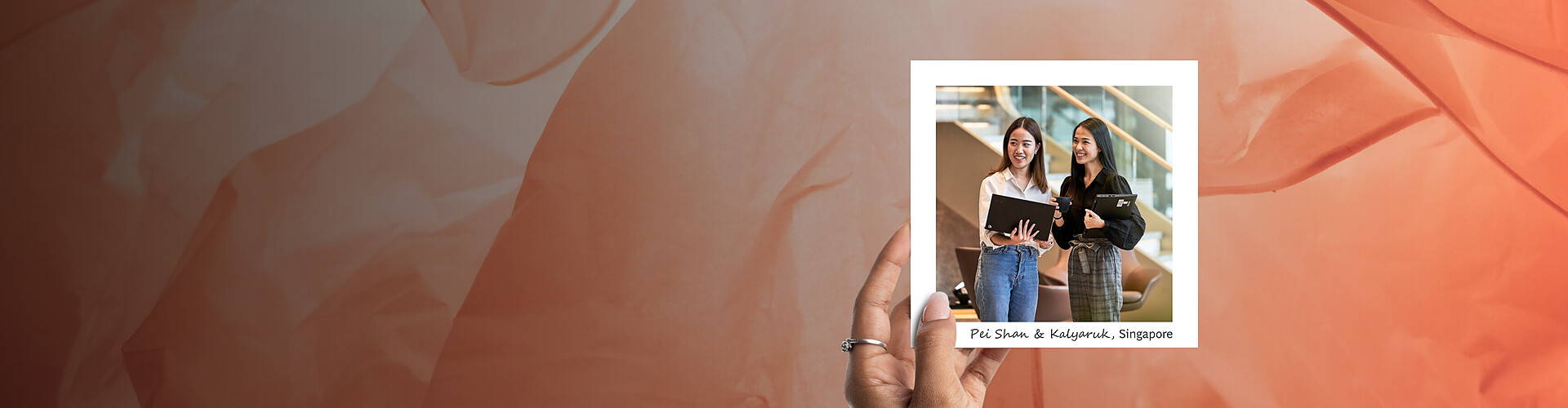  This panoramic banner features a hand holding a photograph against a backdrop of soft, textured orange hues that suggest a warm, welcoming atmosphere. The photograph shows two women, Pei Shan and Kalyaruk, from the Singapore office, walking and engaging in conversation in a modern office environment. They are both dressed in business casual attire and holding tablets, indicating a professional setting. Their expressions are friendly and animated.