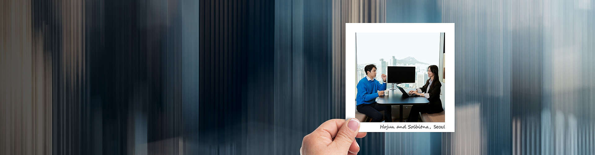 A panoramic banner image featuring a hand holding a photograph against a blurred, abstract blue background. Inside the photograph, two professionals, Hojin and Sobitha, are engaged in a discussion across a table in a well-lit office setting overlooking the cityscape of Seoul. Both are dressed in business attire, and a laptop is visible between them, indicating a work-related meeting.