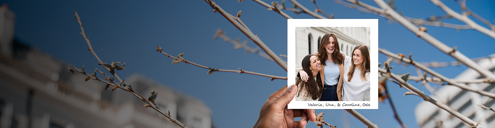 This panoramic banner features a hand holding a photograph against a blurred background of budding branches under a clear blue sky, suggesting the beginning of spring. The photograph captures three women, identified as Valeria, Una, and Caroline from the Oslo office, sharing a joyful moment. They are laughing and embracing each other, clearly enjoying a sunny day outdoors. Their casual attire and genuine expressions convey warmth and camaraderie, emphasizing a close-knit, friendly workplace culture.