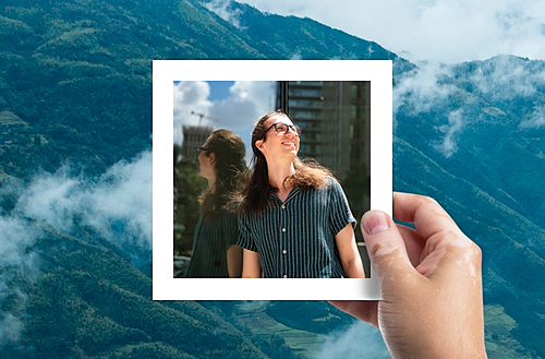 The image features a hand holding a photograph against a scenic backdrop of blue, cloud-wrapped mountains. Inside the photo, a man with sunglasses is smiling broadly, looking upwards as if enjoying a sunny day. He is dressed in a casual striped shirt, and the setting suggests an urban environment with visible building structures. 