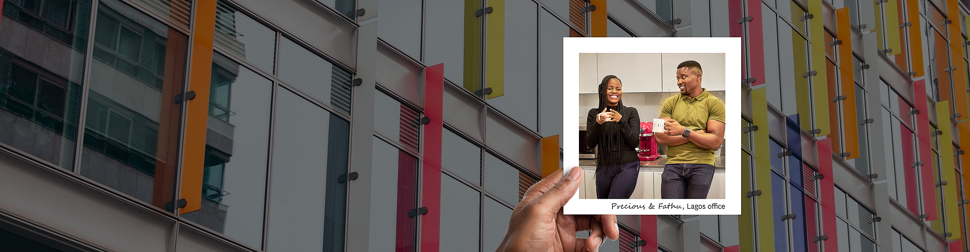 A panoramic banner featuring a hand holding a photograph against a vibrant background of a modern building with colorful, geometric panels. Inside the photo, two colleagues, Precious and Fathu, are shown sharing a light moment in a kitchen at their Lagos office. Precious is smiling and holding a mug, while Fathu is laughing and gesturing with his hand, both exuding a friendly, relaxed office atmosphere. 