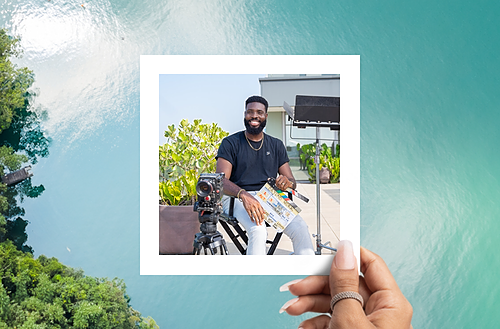 This image shows a hand holding a photograph with a vibrant tropical background featuring clear turquoise water and lush greenery. Inside the photograph, a man is seated outdoors on a rooftop with a camera and equipment set up next to him. He is holding a clapperboard and smiling, suggesting he is either planning or has just completed a project, likely related to filmmaking or photography. 