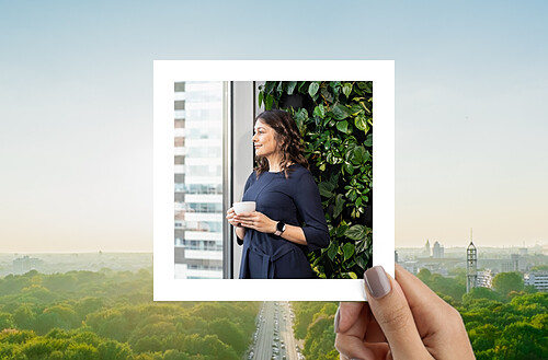 This image depicts a hand holding a photograph in front of a scenic cityscape. The photograph captures a woman standing next to a lush vertical garden inside a modern office building. She is holding a cup, looking out through the large window with a thoughtful expression, possibly enjoying a break or reflecting on her work. The cityscape in the background shows a clear sky and green spaces.