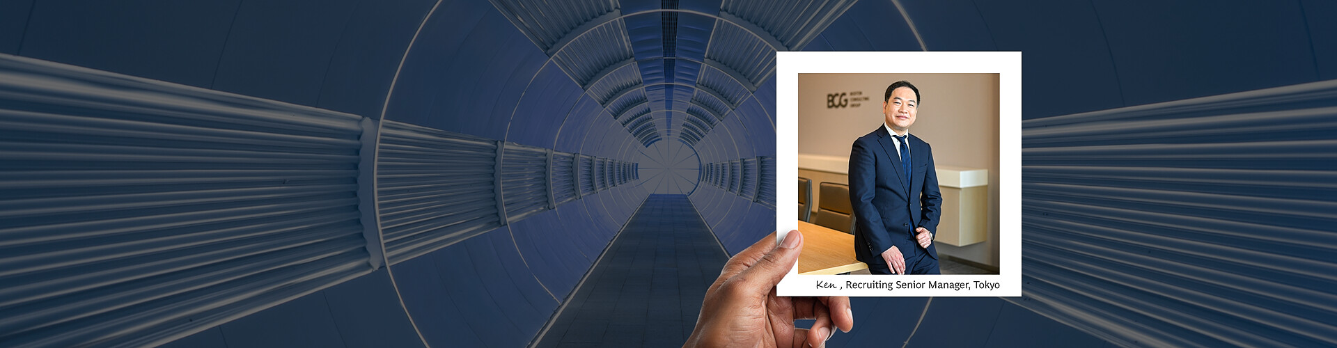 This panoramic banner shows a hand holding a photograph against a visually striking background of a radial blue architectural tunnel. The photograph features Ken, a Recruiting Senior Manager in Tokyo, standing in a modern office environment, dressed in a formal suit and smiling confidently. 