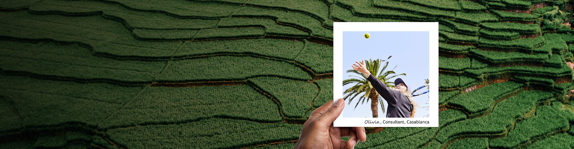 This panoramic banner features a hand holding a photograph against a lush backdrop of terraced tea plantations, vividly showcasing layers of rich greenery. The photograph captures Olivia, a consultant from Casablanca, in action during a tennis match under a clear blue sky, with palm trees in the background. She is reaching upwards to hit a tennis ball, dressed in athletic wear, suggesting a moment of leisure and activity..