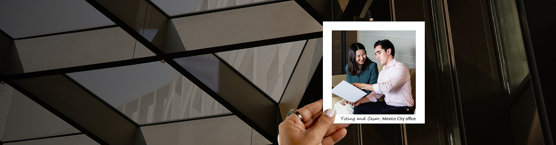 A panoramic banner featuring a hand holding a photograph against an architectural backdrop of dark, angular ceiling panels. Inside the photo, Yiting and Cesar, two colleagues from the Mexico City office, are engaged in a collaborative moment, reviewing a document together. They are smiling and seated closely in a brightly lit office space, suggesting a positive and productive work environment. 