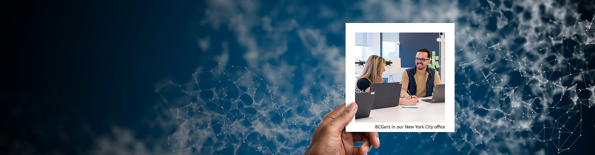 This panoramic banner image features an abstract digital network of interconnected lines and nodes on a dark blue background, symbolizing connectivity and technological integration. A hand is shown holding a photograph of two professionals, a man and a woman, engaged in a discussion at a desk in a modern office setting. The label "BCGers in our New York City office" is included in the photo, indicating the location of the scene. 