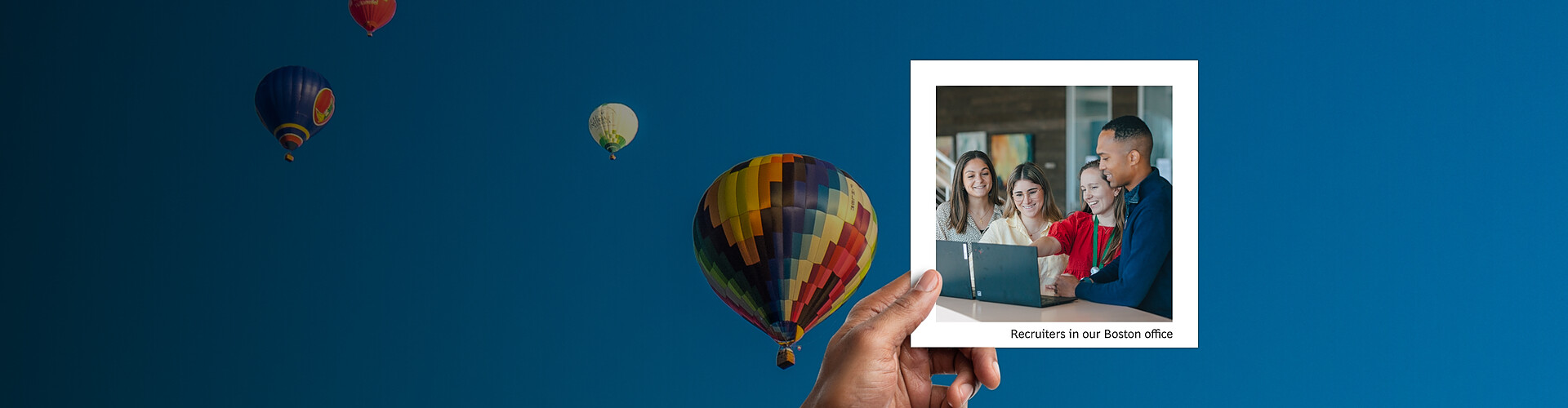 This panoramic banner features a vibrant and clear blue sky as a background, dotted with several colorful hot air balloons in flight, creating a sense of openness and adventure. Overlaid on this sky scene is a smaller photograph of a group of four recruiters, identified as being in the Boston office. They are seen gathered around a laptop, engaging in what appears to be a lively discussion or review of materials. The individuals are diverse and dressed in casual to smart casual office attire, indicating a collaborative and informal office environment. 