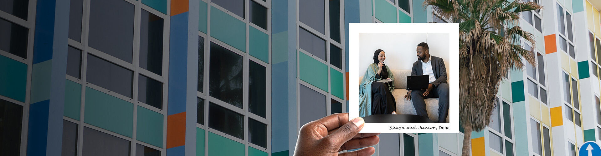 This panoramic banner image features a vibrant and colorful architectural background with a modern building facade decorated in a grid of windows framed in teal, orange, and yellow. A hand is holding a photograph showing two professionals, Shaza and Junior, engaged in a discussion in an office setting in Doha. Shaza, wearing a traditional hijab, is smiling and looking at a smartphone, while Junior is focused on a laptop. 