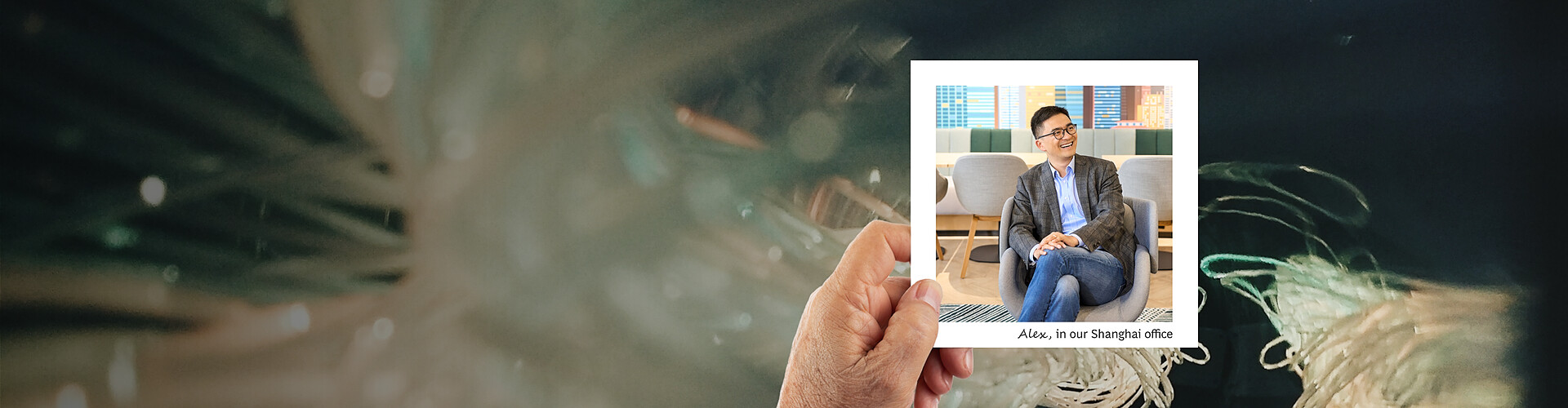 This panoramic banner image features an artistic, blurred background of what appears to be a natural element, possibly plant fronds, in a close-up view. A hand is holding a photograph of a man named Alex, labeled as being in the Shanghai office. In the photo, Alex is seated comfortably in a modern office lounge, dressed in a business casual blazer and jeans, laughing and appearing very relaxed. 