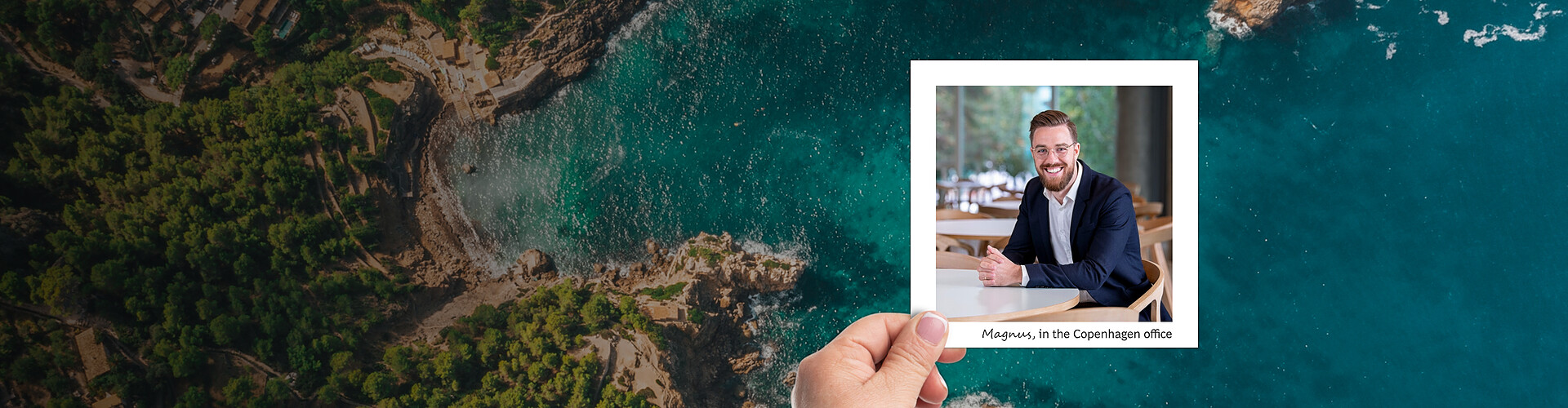  This is a panoramic banner image, where the main focus is a beautiful coastal scene overlaid by a smaller portrait of a man named Magnus, who is seated in an office setting in Copenhagen. The coastal scene features a bird’s-eye view of a rugged shoreline with turquoise waters, dense greenery, and a serpentine road weaving through the landscape. The portrait of Magnus shows him smiling and dressed in business attire, sitting at a table, suggesting a professional environment. His name and office location are indicated in the text accompanying his image.