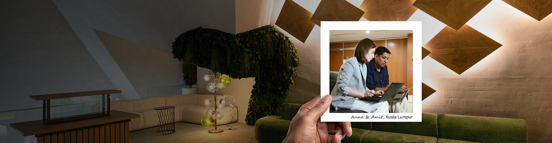 This panoramic image features a hand holding a photograph against a stylish office interior. The office backdrop includes angular geometric patterns on the wall, a plush green sofa, and an artistic tree sculpture adorned with lights, creating a warm and inviting space. In the photograph, a man and a woman, identified as Anna and Amit from Kuala Lumpur, are focused on a laptop screen, engaged in a professional discussion.
