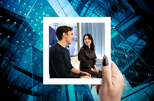  A creative image featuring a hand holding a Polaroid photo against a futuristic blue glass building background. Inside the photo, a young man and woman are engaging in a conversation in a modern office setting, with the woman smiling and the man facing her with an attentive expression.