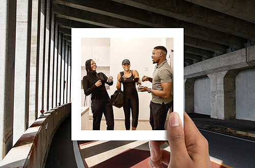 A photo within a photo concept where a hand holds up a Polaroid image of three young, fit people—two women and one man—chatting and laughing together in a bright, modern gym environment. The backdrop outside the Polaroid is an urban scene with a curved road under a concrete bridge, contrasting the enclosed gym setting with the openness of the urban environment.