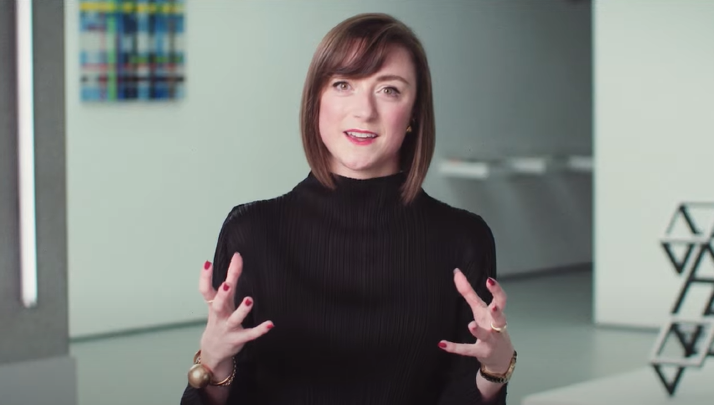 The thumbnail photo features a woman with shoulder-length brown hair, wearing a black top, and gesturing with her hands as she speaks. She has a friendly expression and is situated in a modern, minimalist indoor setting with abstract art and geometric decor visible in the background. A white play button icon is centered on the image, indicating that the thumbnail is for a video. 