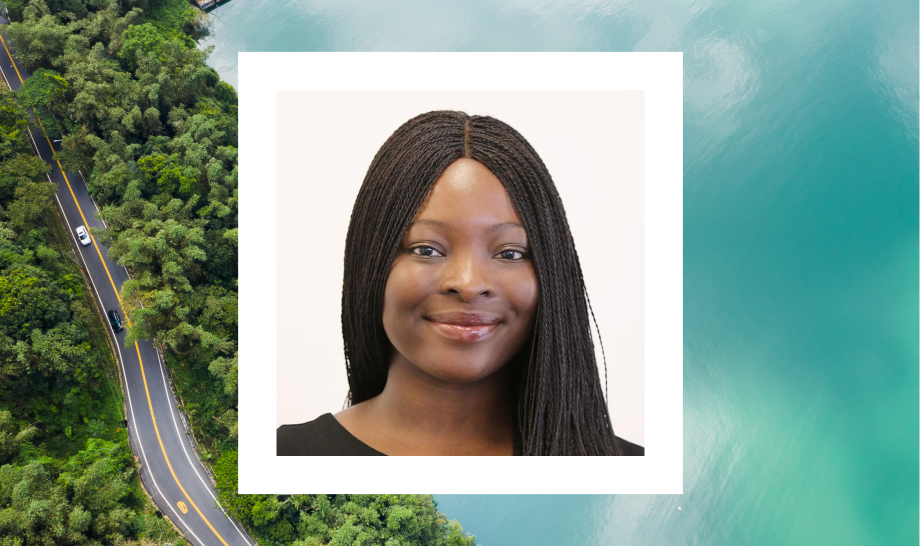 A photo of a woman with dark skin and long braided hair that is parted in the middle is in a white frame. She is smiling gently and is wearing a black top. The background features a scenic view of a winding road through lush green trees on one side and a body of water on the other, creating a tranquil and picturesque atmosphere.