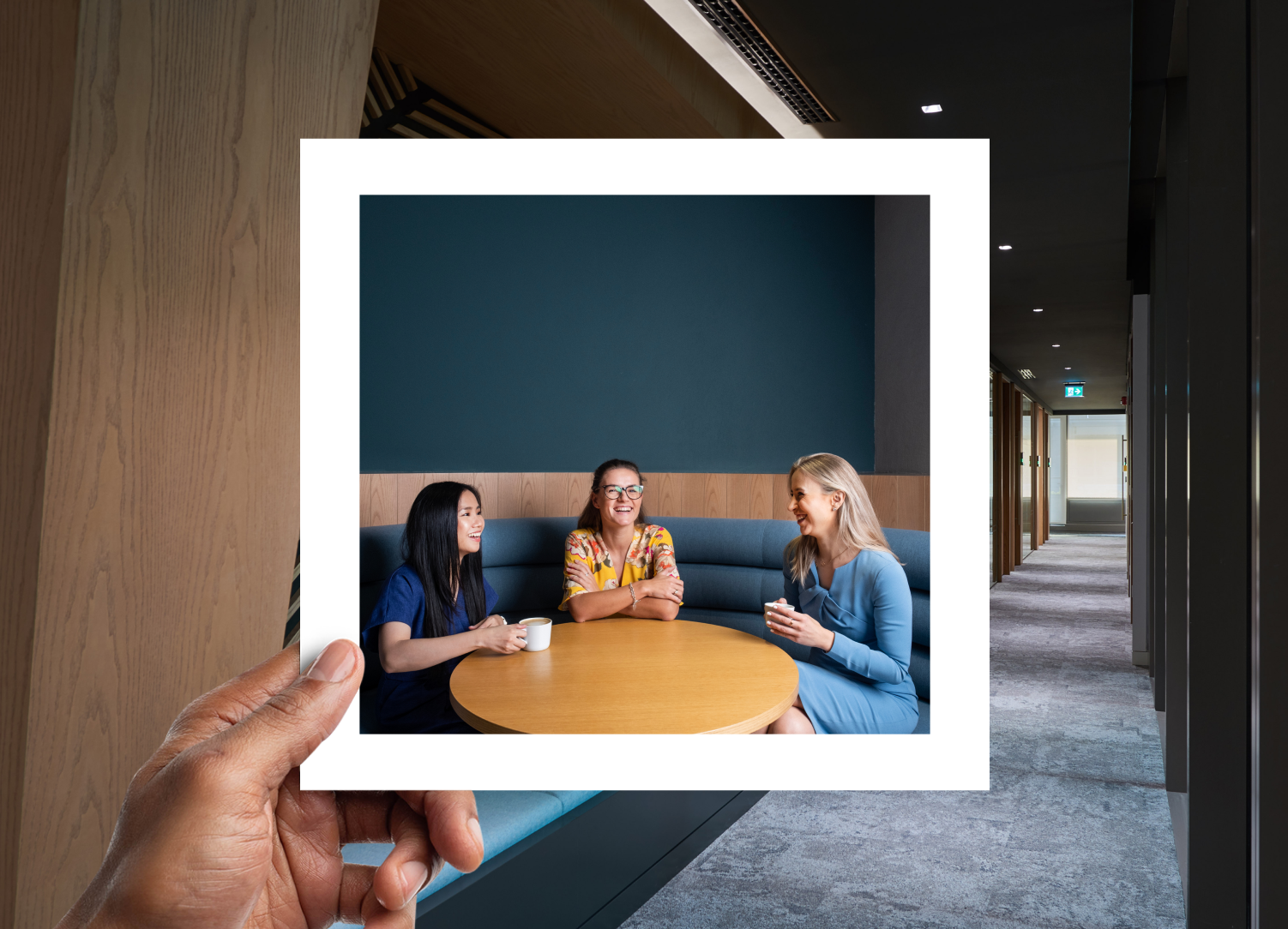  A modern office setting with three women sitting around a circular table, smiling and having a conversation over coffee, framed within a polaroid-style overlay.