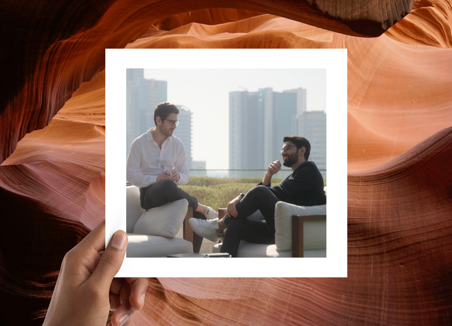 Two men sitting on a rooftop lounge, enjoying a conversation with city buildings visible in the background, framed in a polaroid-style overlay with a natural sandstone texture as the border.