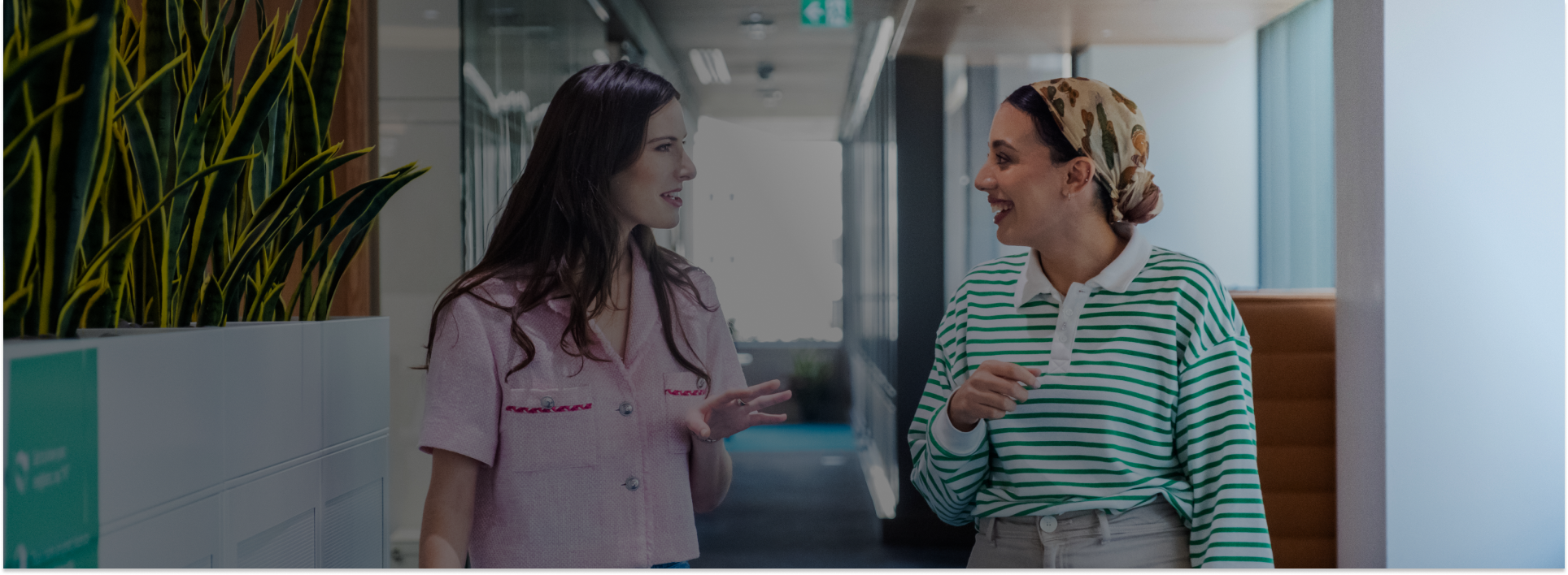 two women walking and engaging in a conversation inside an office hallway with greenery and modern interiors.