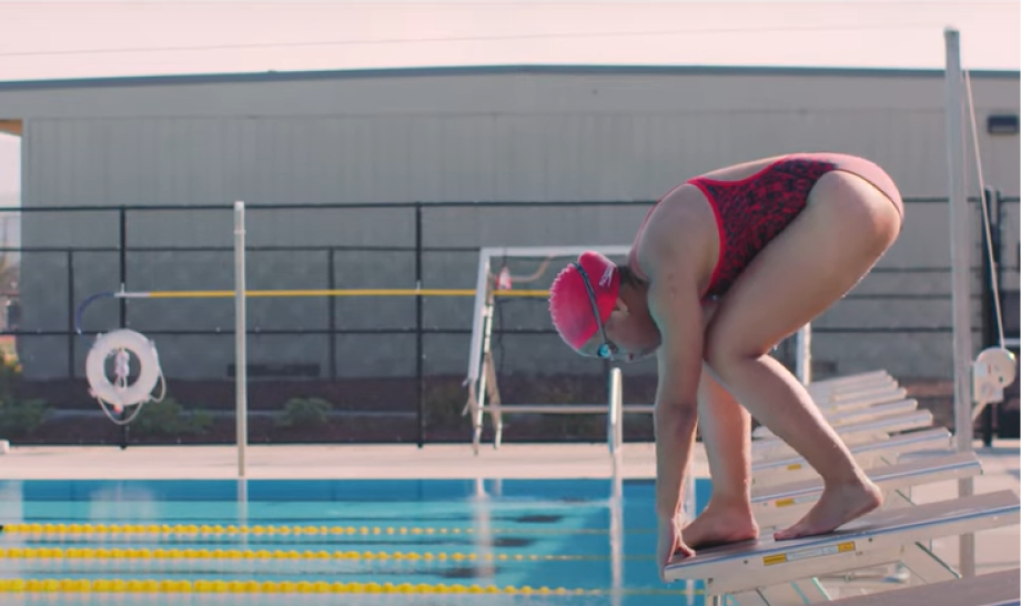 A swimmer is in a starting position on a diving platform at the edge of a pool. She is wearing a red swimsuit with a pattern, a pink swim cap, and goggles. The pool has clear blue water with yellow lane dividers and is situated outdoors with a building and fencing in the background. The swimmer is bent over, poised to dive into the water.