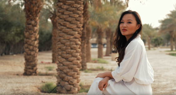 A woman in a white outfit sits calmly on a bench in a palm tree-lined park. She gazes towards the camera with a serene expression, her long dark hair cascading over her shoulders. The background features a path winding through the park, dotted with palm trees and soft sunlight filtering through the leaves, creating a tranquil and warm atmosphere.