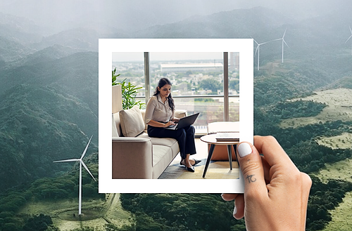 The image features a hand holding a photograph against a scenic backdrop of green, rolling hills dotted with wind turbines, emphasizing sustainability. Within the photograph, a professional woman sits comfortably on a sofa, working on her laptop next to a window that offers a view of a lush, urban landscape. 