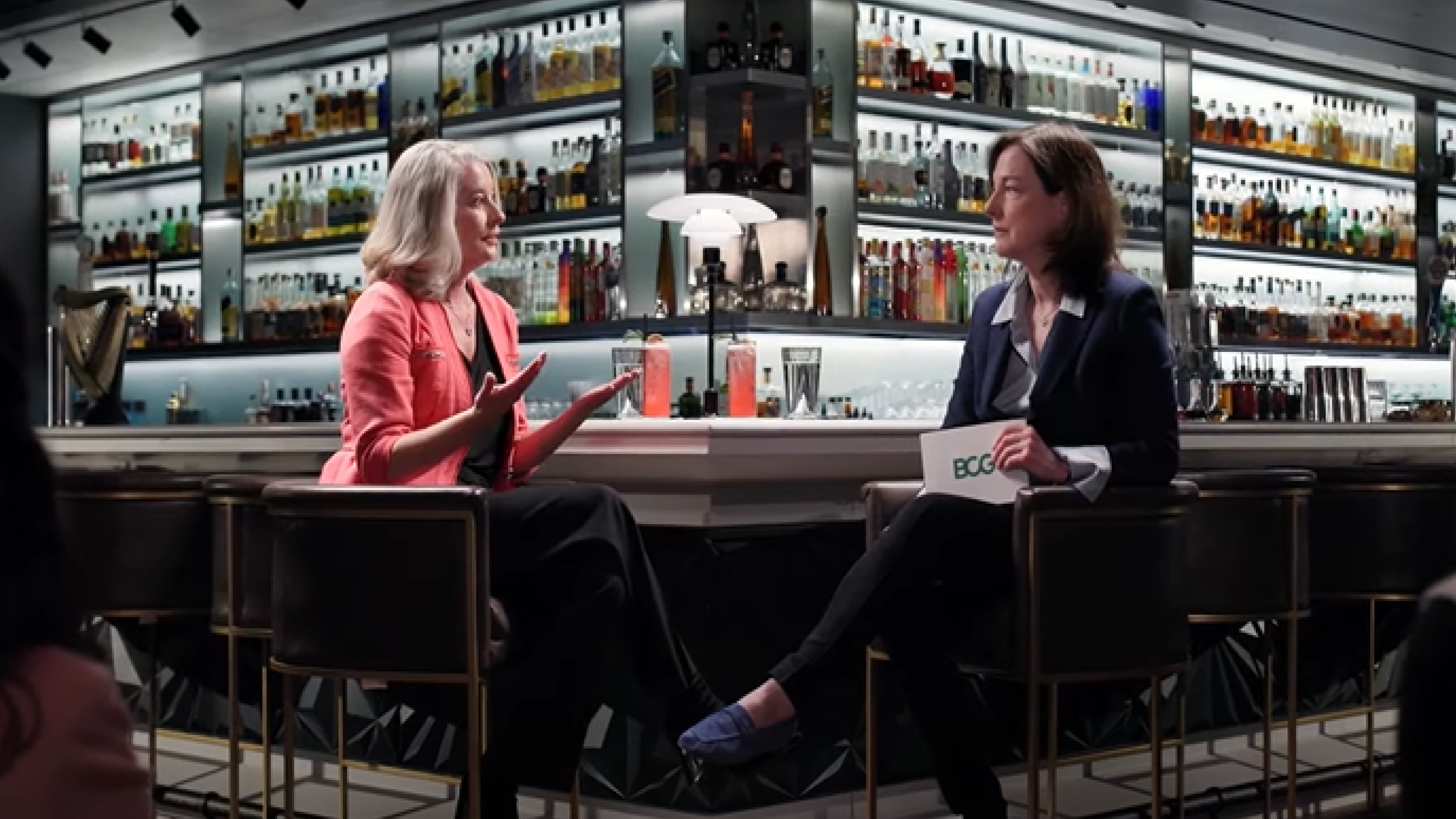 Two women are seated at a bar engaged in conversation. The woman on the left, wearing a coral blazer, is gesturing with her hands while speaking. The woman on the right, in a navy suit, holds a card with "BCG" on it, listening attentively. The background features shelves filled with various bottles, suggesting a well-stocked bar setting.